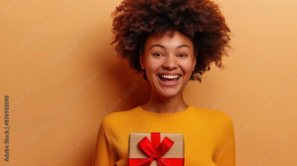 Smiling woman with curly hair holding a wrapped gift box in front of an orange background, conveying joy and surprise. Smiling woman with curly hair holding a wrapped gift box in front of an orange background, conveying joy and surprise.