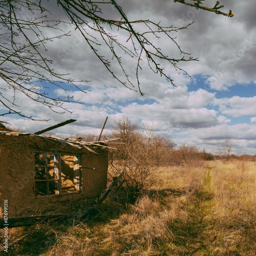 an old broken clay house without a roof, with a wooden window, around which bare trees and bushes grow, against a background of a blue sky with white clouds