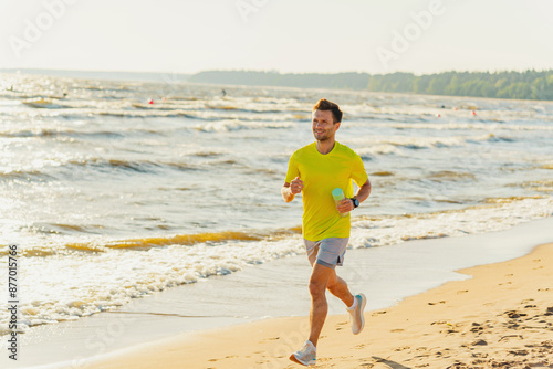 A man in a vibrant yellow shirt jogs along the beach, holding a cup, waves shimmering in the sunlight.