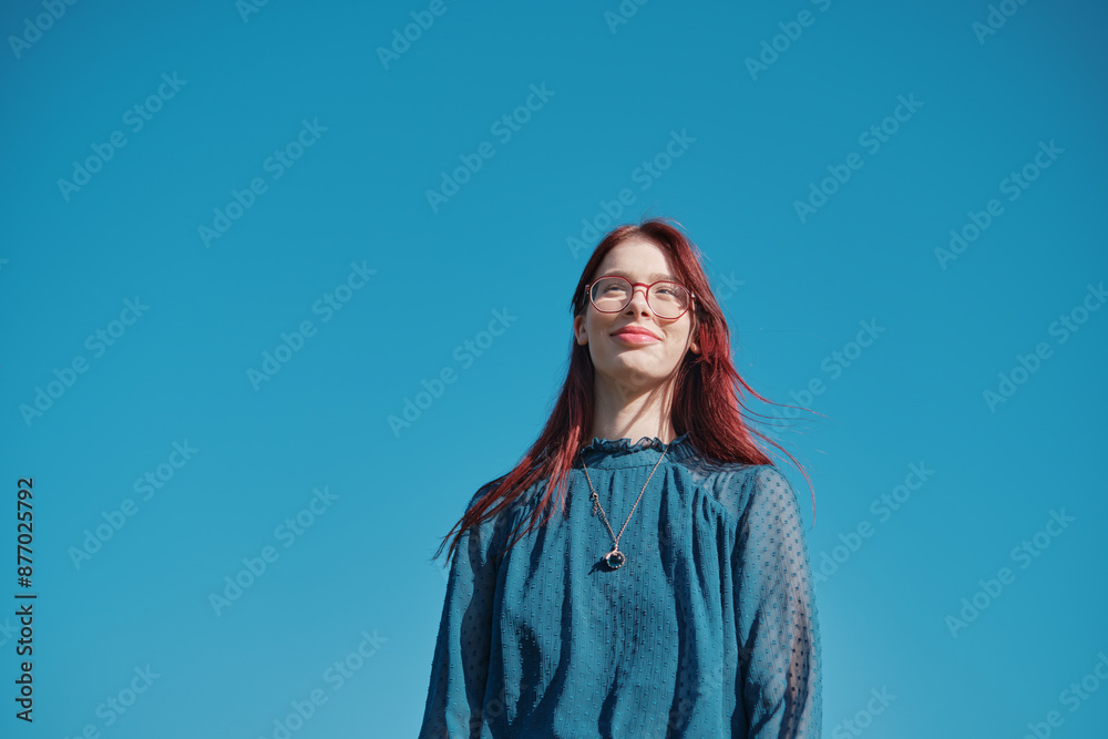 Teen girl in blue blouse and coral skirt standing on green grass with one hand in pocket against clear blue sky, low angle shot Teen girl in blue blouse and coral skirt standing on green grass with one hand in pocket against clear blue sky, low angle shot