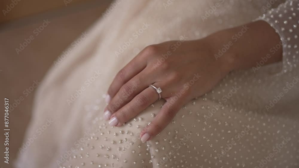 Close-up of a bride's hand resting on a beaded wedding dress, showing a diamond engagement ring. Close-up of a bride's hand resting on a beaded wedding dress, showing a diamond engagement ring.