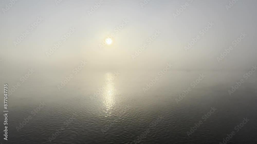 A wide static shot of sunrise amidst morning fog taken over a flowing water body with sun's reflection