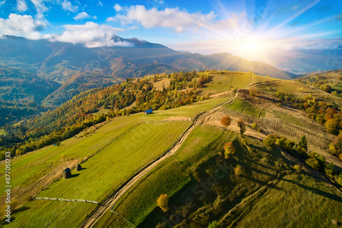 Aerial view of mountain road at sunset in autumn. Top view from drone of road in hills. Beautiful landscape with roadway, trees, green meadows, sky with golden sunlight in fall.