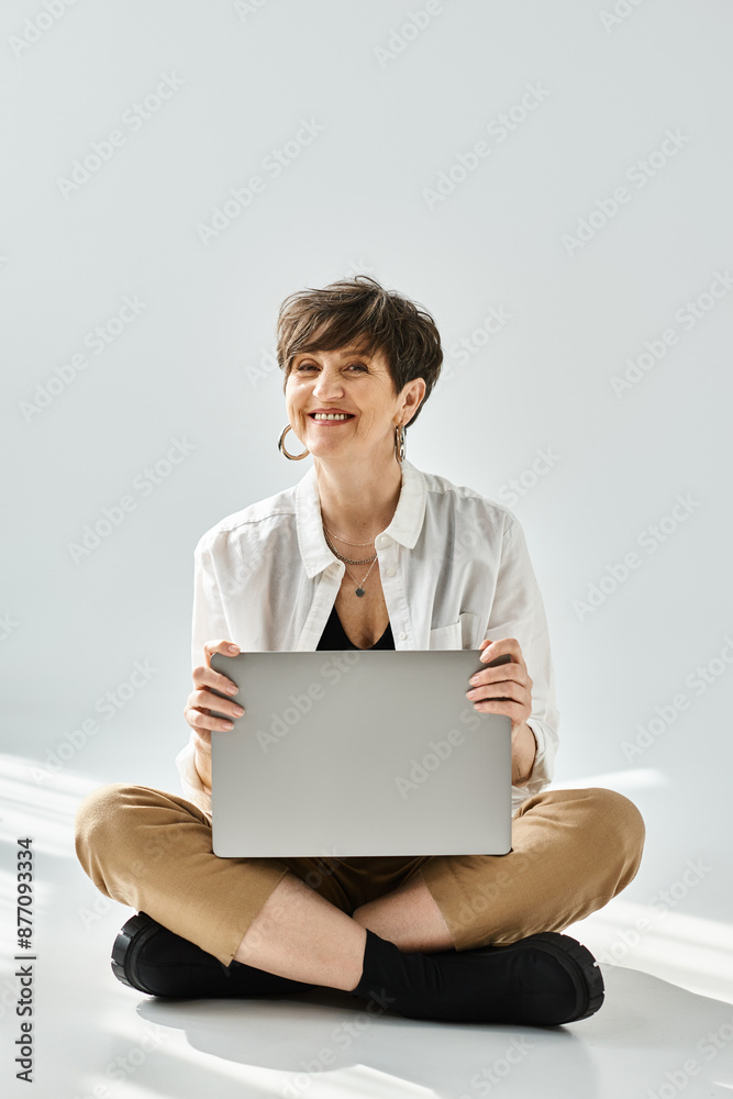 A stylishly dressed middle-aged woman with short hair is sitting on the floor, working on a laptop in a studio setting. A stylishly dressed middle-aged woman with short hair is sitting on the floor, working on a laptop in a studio setting.