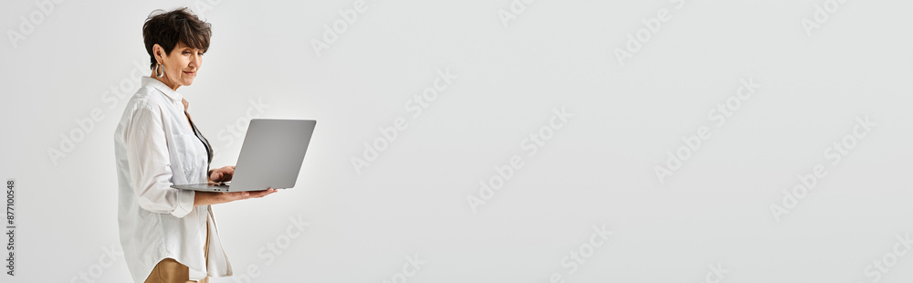 A middle-aged woman stands confidently holding a laptop in a stylish studio setting.