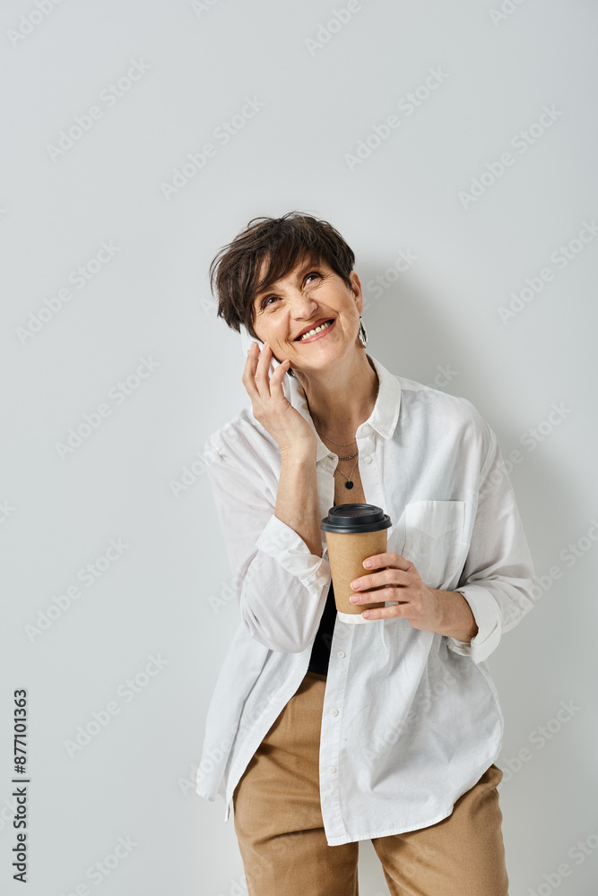 A middle-aged woman with short hair, dressed stylishly, multitasking by holding a cup of coffee and chatting on her cellphone. A middle-aged woman with short hair, dressed stylishly, multitasking by holding a cup of coffee and chatting on her cellphone.