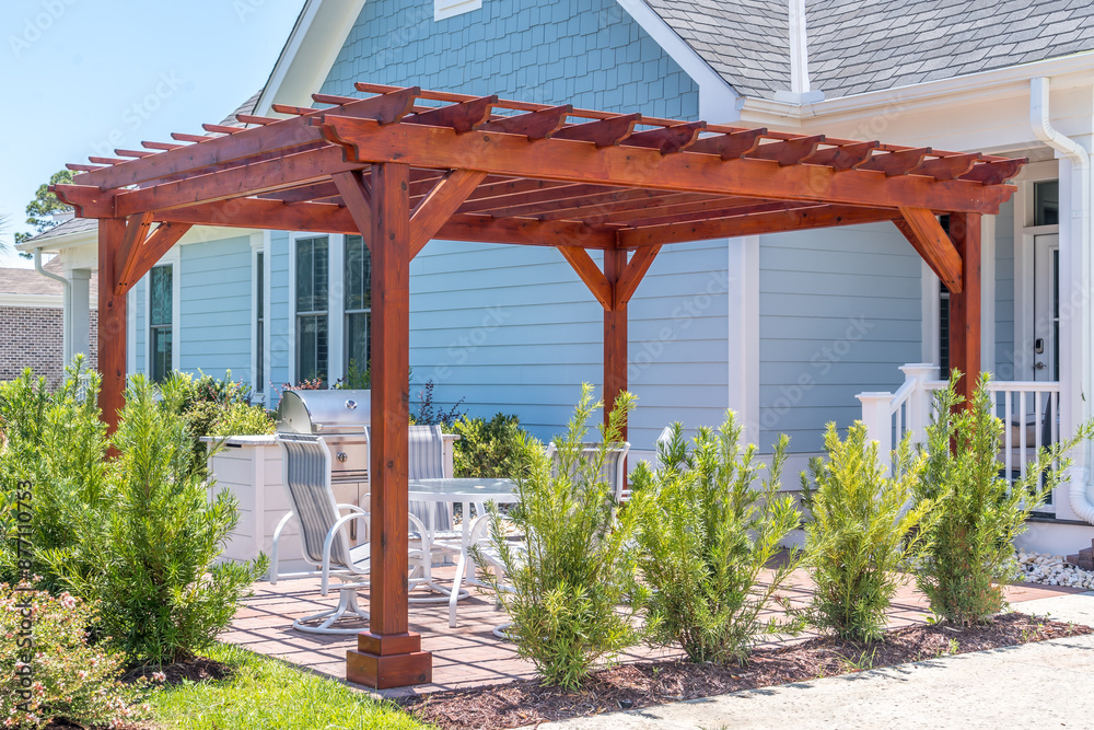 Trendy outdoor patio pergola shade structure made of dark wood, awning and patio roof, white dining table, white chairs, metal grill surrounded by landscaping in a luxury American neighborhood