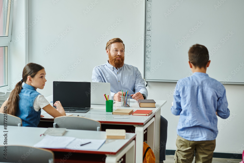 A man sits at a desk in front of a group of children, engaging them in a lively classroom setting. A man sits at a desk in front of a group of children, engaging them in a lively classroom setting.