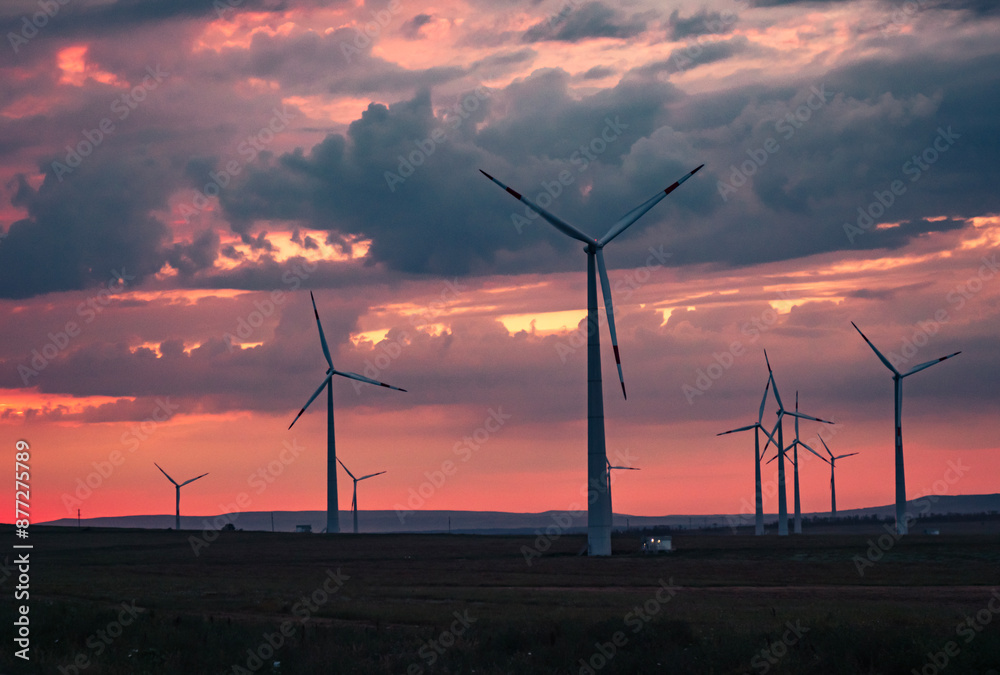 Obraz premium Multiple wind turbines silhouetted against a vibrant sunset sky. The turbines stand tall against the backdrop of orange and red clouds.