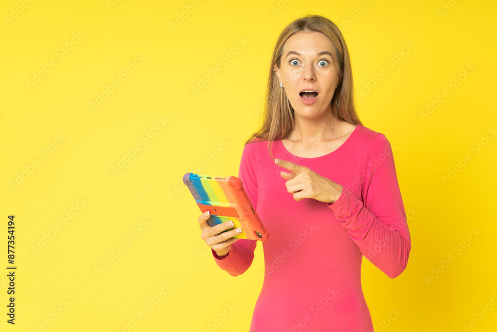 Shocked excited young blond woman with tablet pc isolated on yellow background. Surprised woman with open mouth in red fitted dress pointing to tablet pc holding in hand Shocked excited young blond woman with tablet pc isolated on yellow background. Surprised woman with open mouth in red fitted dress pointing to tablet pc holding in hand
