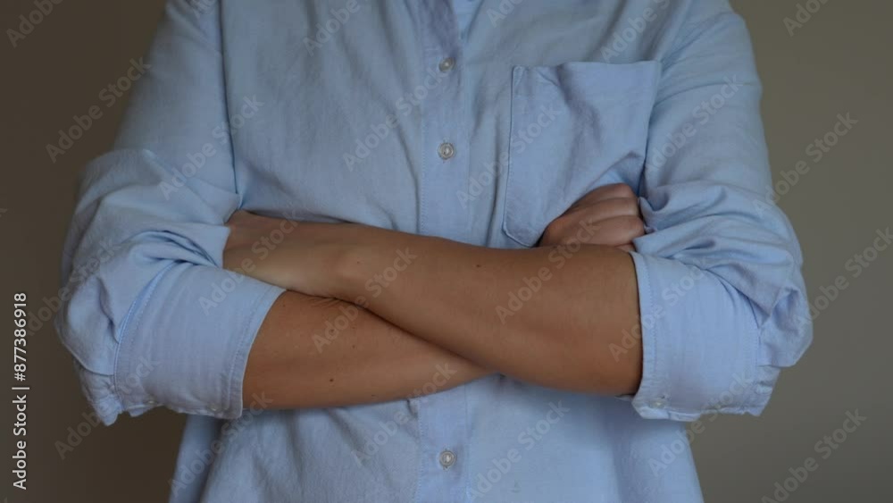 A cropped shot of a young woman in a blue shirt crossing her arms on her stomach on a grey background. Closed pose A cropped shot of a young woman in a blue shirt crossing her arms on her stomach on a grey background. Closed pose