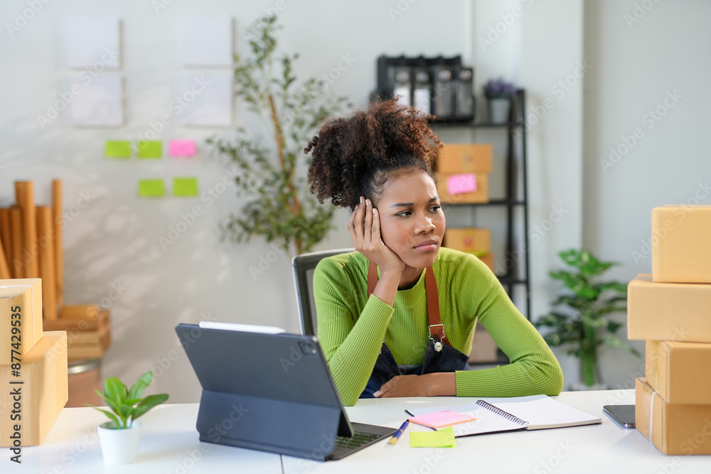 Stressed young entrepreneur sitting at her desk with paperwork and boxes, feeling overwhelmed Stressed young entrepreneur sitting at her desk with paperwork and boxes, feeling overwhelmed