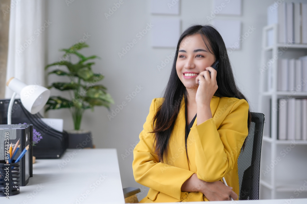 Smiling young businesswoman in a modern office, wearing a yellow blazer, talking on her phone at her desk Smiling young businesswoman in a modern office, wearing a yellow blazer, talking on her phone at her desk