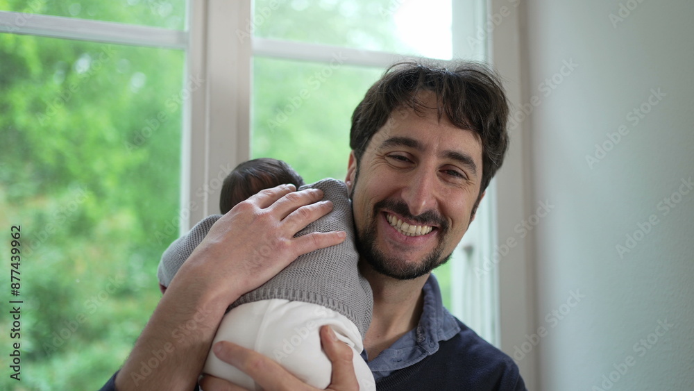 Newborn baby resting on the father’s shoulder, dressed in a cozy knitted sweater. The father turns body towards window, highlighting the comforting and secure embrace between them