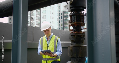 A Engineer man looking inspecting maintenance insulated pipelines valve pump control on the roof at an industrial site. He is wearing a hard hat and safety vest