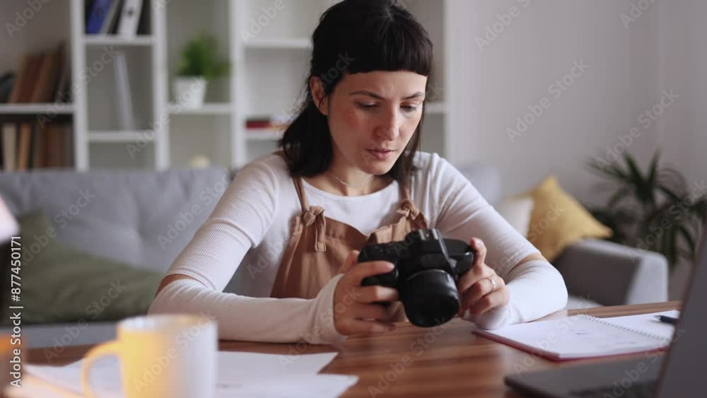 Portrait of pretty young caucasian woman photographer hold digital camera looking at screen choosing photos for editing while sitting in front of laptop computer at home workplace Portrait of pretty young caucasian woman photographer hold digital camera looking at screen choosing photos for editing while sitting in front of laptop computer at home workplace