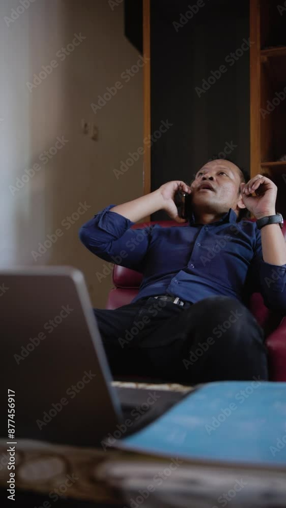 Asian Indonesian male working answering on phone call. Man talking on smartphone sitting on sofa at remote workplace. Business man calling phone at office. Asian Indonesian male working answering on phone call. Man talking on smartphone sitting on sofa at remote workplace. Business man calling phone at office.