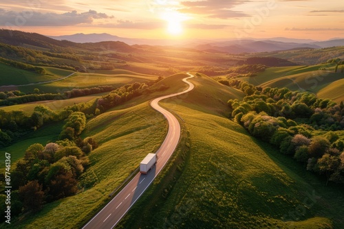 Amidst the serene countryside, a truck travels along a winding road under the warm hues of the setting sun. Experience the peaceful journey through.