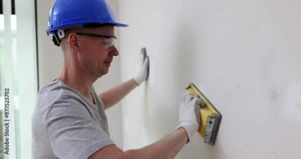 Foreman in blue helmet processes white walls in living room with tool. Focused worker in goggles and protective gloves does renovation slow motion Foreman in blue helmet processes white walls in living room with tool. Focused worker in goggles and protective gloves does renovation slow motion
