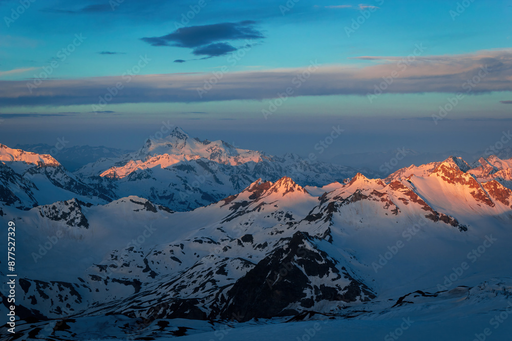 Obraz premium Golden sunset lights snowy peaks of Caucasus Range. Majestic mountain landscape glow at dusk. Clear blue sky, white clouds. Nature beauty, travel destination for hikers, photographers.