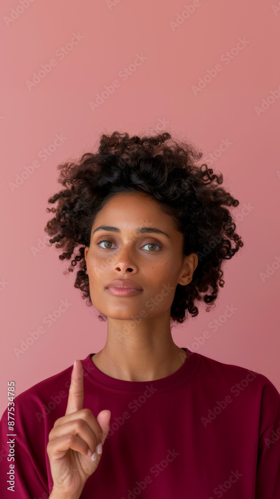 Portrait of an adult African American woman in a burgundy t-shirt looking into the frame and holding her raised hand with her index finger up, pink solid background