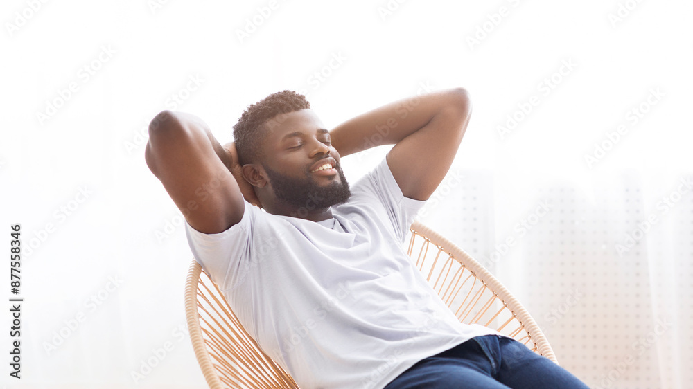 A Black man with a beard, wearing a white t-shirt and blue jeans, sits relaxed in a wicker chair with his arms behind his head. He is looking off to the side, with a serene expression on his face A Black man with a beard, wearing a white t-shirt and blue jeans, sits relaxed in a wicker chair with his arms behind his head. He is looking off to the side, with a serene expression on his face