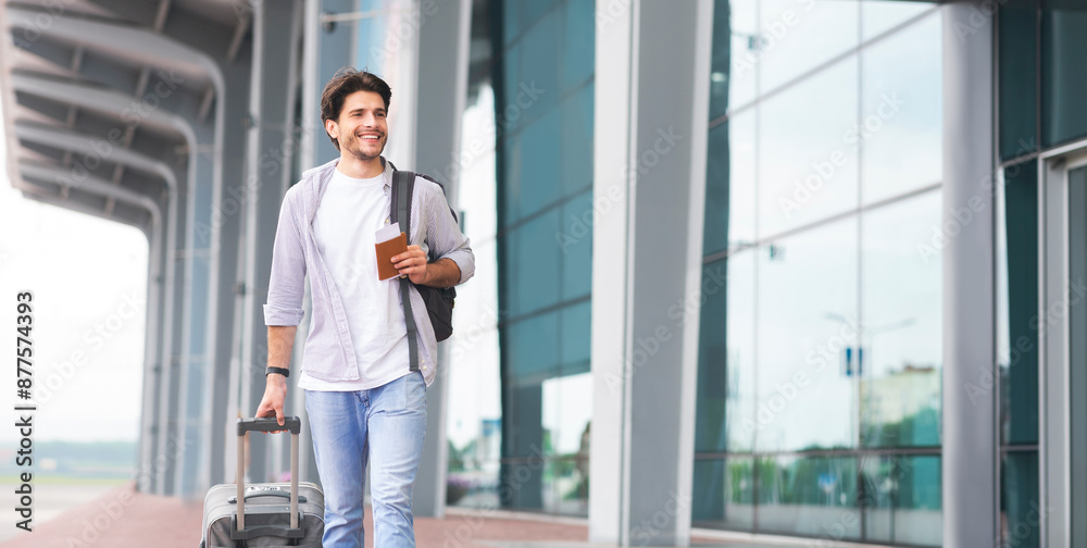 Traveling concept. Happy man in casual clothes walking outdoors near airport terminal with luggage, passport and tickets, ready for trip, free space Traveling concept. Happy man in casual clothes walking outdoors near airport terminal with luggage, passport and tickets, ready for trip, free space
