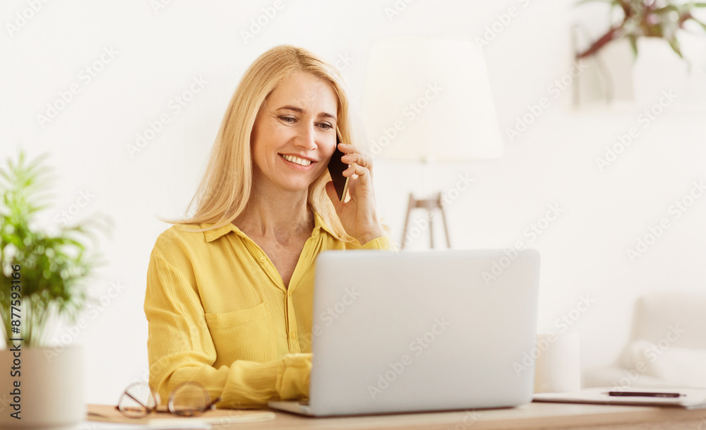A woman multitasks with a smile, working on her bright home offices laptop while chatting on her phone, with a plant in the background. A woman multitasks with a smile, working on her bright home offices laptop while chatting on her phone, with a plant in the background.
