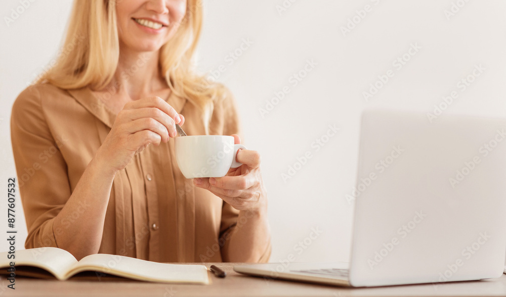 A woman sits at a desk, stirring coffee in a white mug while working on a laptop. A woman sits at a desk, stirring coffee in a white mug while working on a laptop.