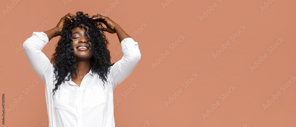 This image shows a smiling, Black woman with long, curly hair against a light brown background. She is wearing a white, long-sleeve shirt and is touching her hair with both hands. This image shows a smiling, Black woman with long, curly hair against a light brown background. She is wearing a white, long-sleeve shirt and is touching her hair with both hands.