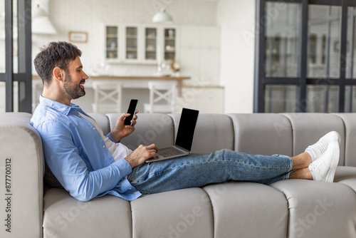 A man is sitting on a gray couch in a modern living room, using his laptop and looking at his smartphone. He is wearing a light blue shirt and blue jeans, and his feet are up on the couch.
