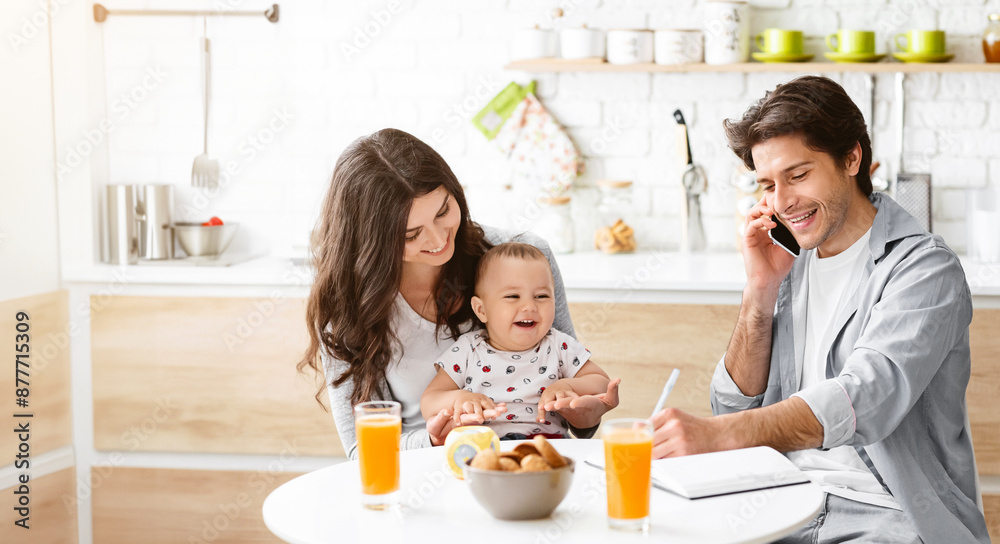 A happy family enjoys a casual breakfast together in their kitchen. A young woman holds her baby in her lap, and a man sits at the table, smiling while talking on the phone. A happy family enjoys a casual breakfast together in their kitchen. A young woman holds her baby in her lap, and a man sits at the table, smiling while talking on the phone.