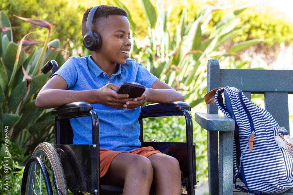Obraz premium Joyful African American boy in wheelchair with headphones, smartphone in garden