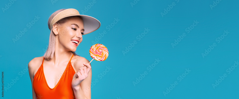 A young woman with blonde hair is smiling at the camera. She is wearing a red swimsuit and a white visor hat, and is holding a colorful lollipop in her hand. The background is a solid blue color. A young woman with blonde hair is smiling at the camera. She is wearing a red swimsuit and a white visor hat, and is holding a colorful lollipop in her hand. The background is a solid blue color.