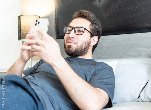 Joven apuesto de 20 años acostado en su habitación. Hombre con anteojos usando el teléfono celular 