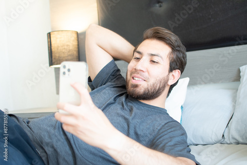 Joven de bella sonrisa usa su teléfono celular. Hombre apuesto con barba acostado en su habitación 