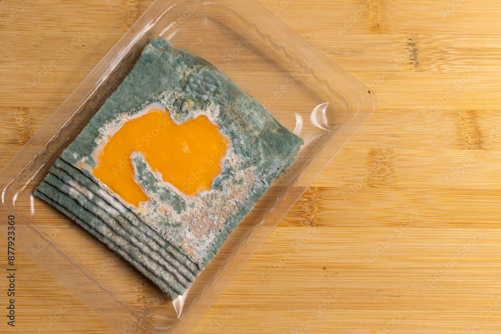 Slices of moldy yellow cheese arranged on a bamboo cutting board. Green mold spores on food. Expired wasted food
