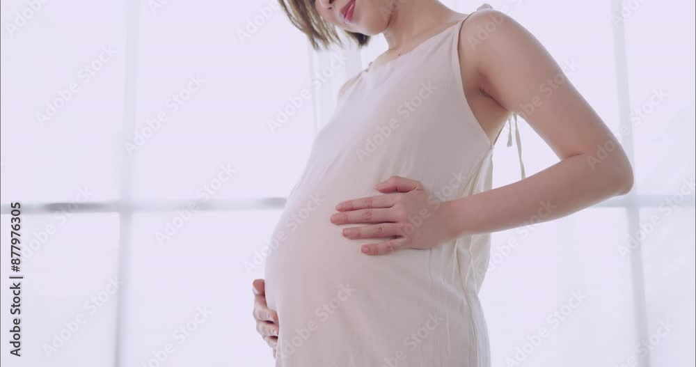 Happy Asian pregnant woman lovingly touching her unborn baby while standing by the window at home.