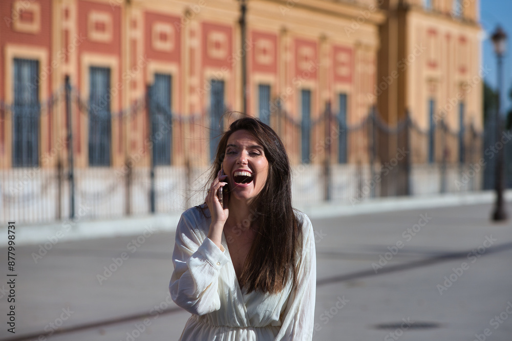 Fototapeta premium Young and beautiful Latin woman talking on mobile phone is having fun and is happy and smiling loudly. The woman is on the street and walks through the city. Concept communications.