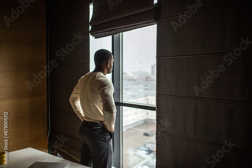 Serious dark skinned businessman, feeling worried about new business project, dressed in knitted white pullover, spends free time at office, looking at cityscape through pamoramic window. photo