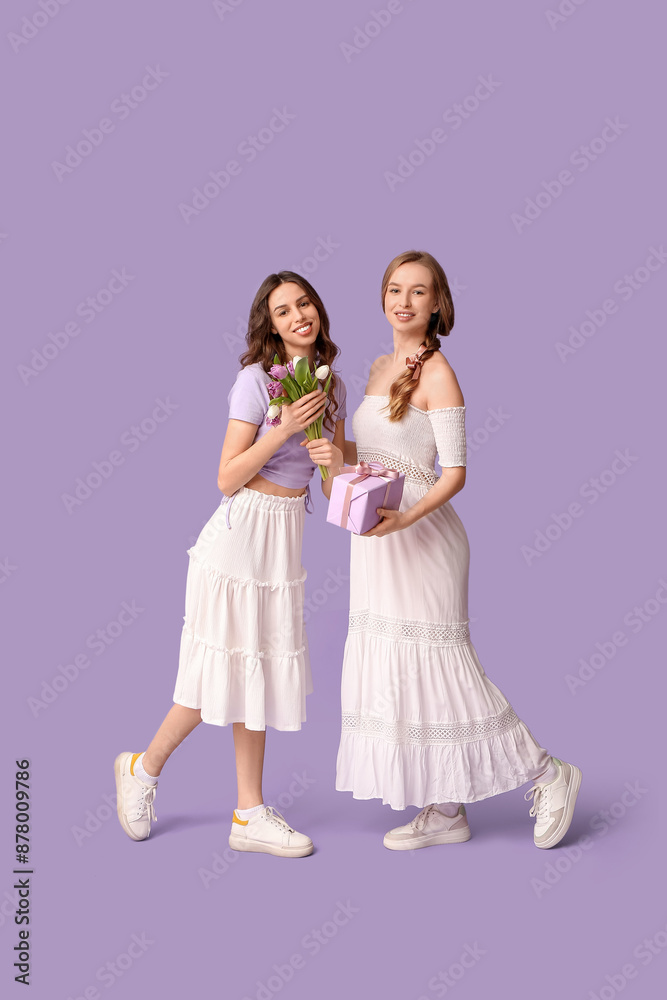 Beautiful young sisters with gift box and tulips on lilac background