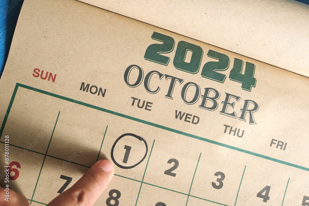 Hand pointing on October 1 2024 date on calendar flat lay in blue background. First day of the month reminder.