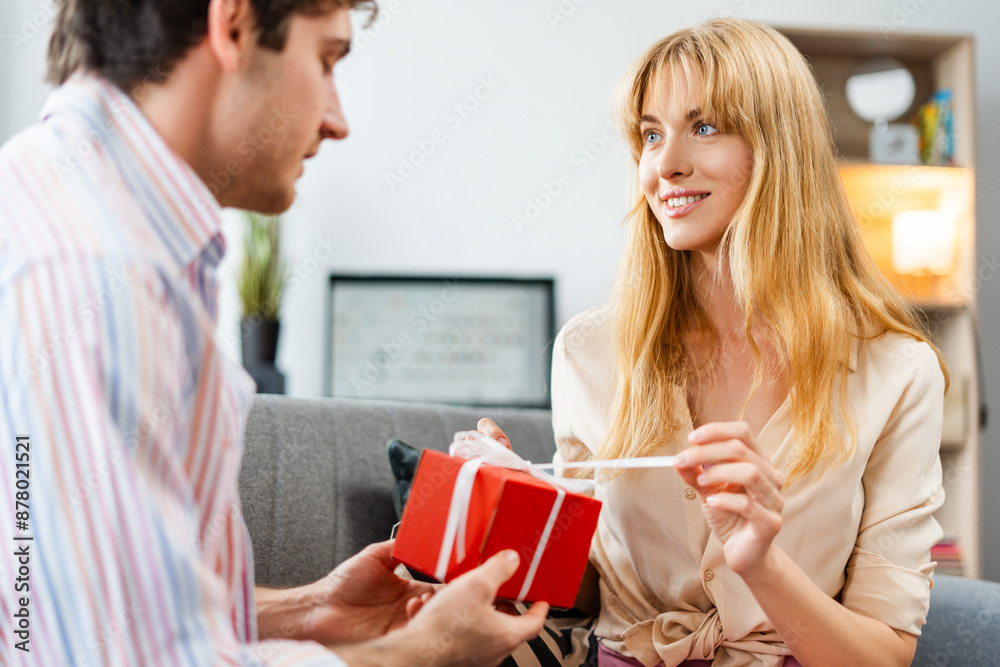 Young woman opening gift given by boyfriend at home, sitting on sofa in living room Young woman opening gift given by boyfriend at home, sitting on sofa in living room