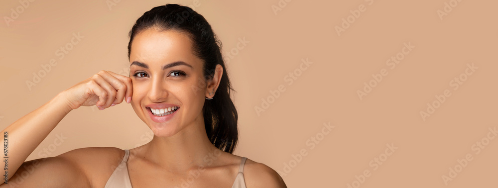 Obraz premium Anti-aging treatment in cosmetology. Smiling attractive millennial middle eastern woman touching her cheek, demonstrating youthful smooth elastic skin, posing on beige studio background, copy space