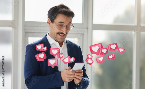 A man wearing a blue suit and glasses smiles as he looks at his smartphone, surrounded by animated pink hearts symbolizing likes or positive feedback photo