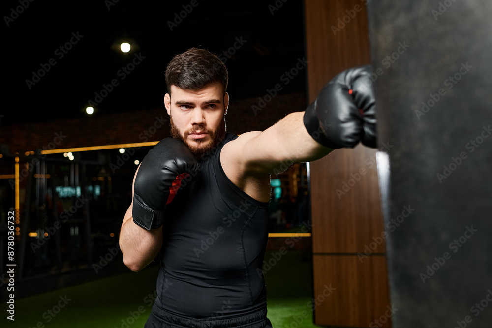 A bearded man in a black shirt trains in a gym, throwing punches at a heavy bag with black boxing gloves.