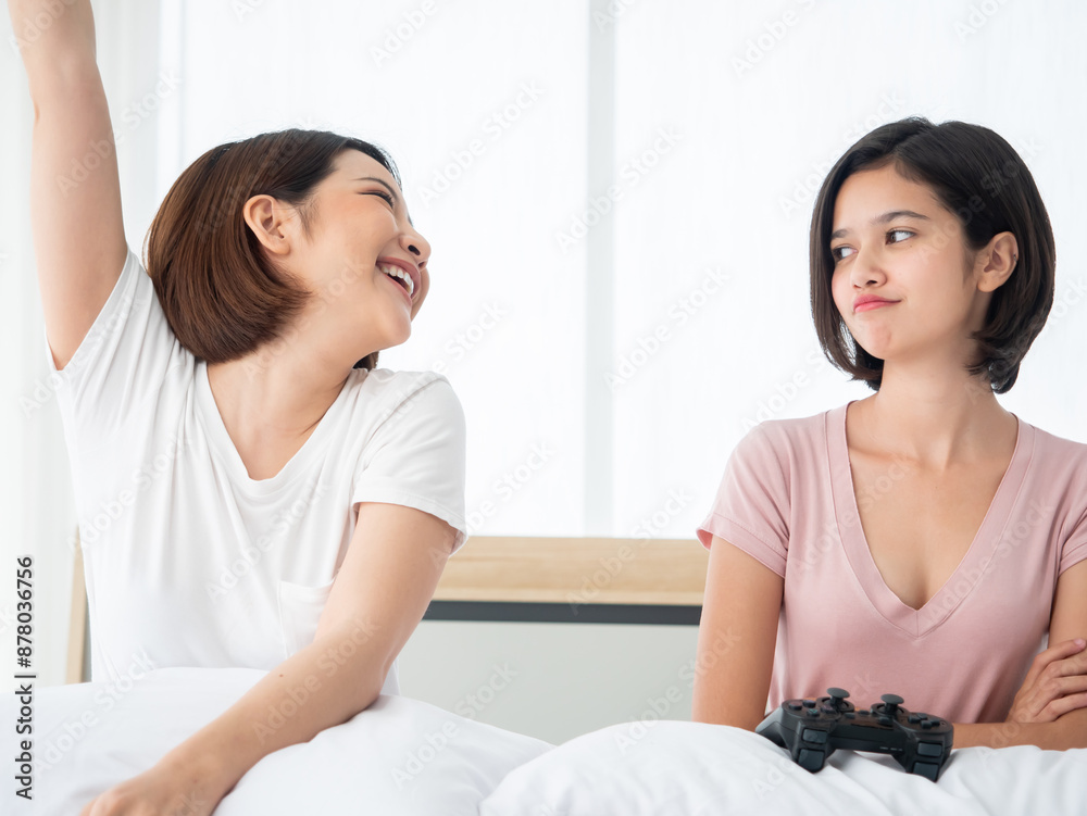 Asian couple playful female sitting on a bed, one celebrating with winner joy raise arm and the loser looking disappointed holding a video game controller Asian couple playful female sitting on a bed, one celebrating with winner joy raise arm and the loser looking disappointed holding a video game controller