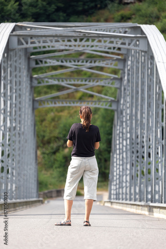 Obraz na plátně Woman walking away on a sturdy, grey metal bridge surrounded by green trees and bushy vegetation, indicating a peaceful and serene outdoor environment