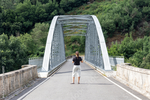 Fényképezés Woman walking away on a sturdy, grey metal bridge surrounded by green trees and bushy vegetation, indicating a peaceful and serene outdoor environment