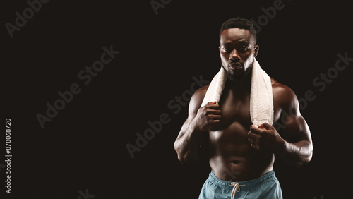 A shirtless, muscular man stands against a black background, holding a towel around his neck, looking intense.